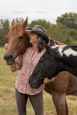 Someone in a cowboy hat and peach shirt embraces two horses - one chestnut brown and one black - in a grassy field.