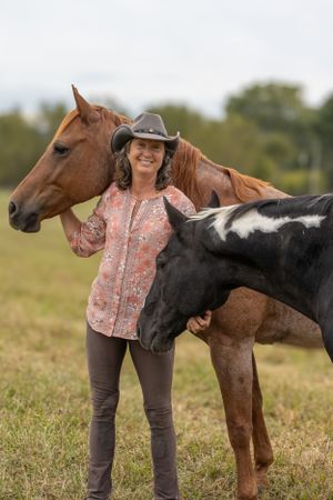 A person poses with two horses - one chestnut brown and one black and white pinto - in a grassy field.