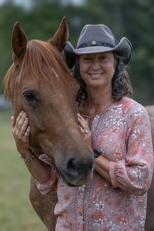 A beautiful chestnut horse and a rider in a floral top and gray cowboy hat share a tender moment outdoors.