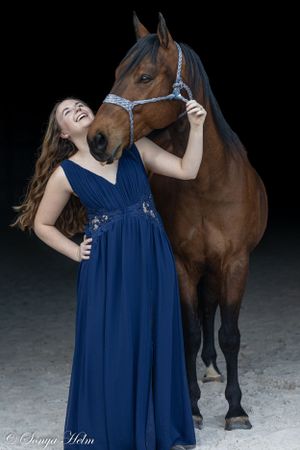 Person in long navy dress shares joyful moment with bay horse against dark backdrop.