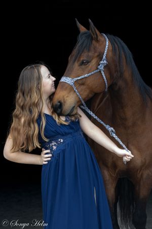 Person in blue dress shares intimate moment with bay horse against black backdrop.