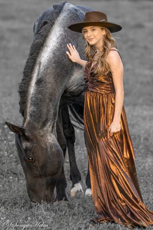 A selective color photo of a grazing horse with someone in a brown dress touching its back.