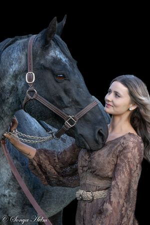 A black horse and companion in brown dress stand together in a dramatic dark portrait setting.