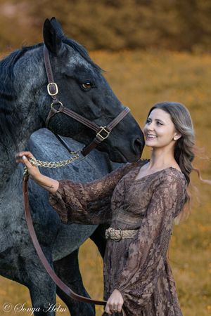 A majestic black horse shares a tender moment with someone wearing a flowing brown vintage dress outdoors.