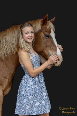 A person in a blue floral dress poses with a chestnut horse with a white blaze against a black background.