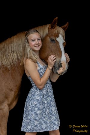 A person wearing a blue patterned dress stands next to a brown horse with a white face marking.