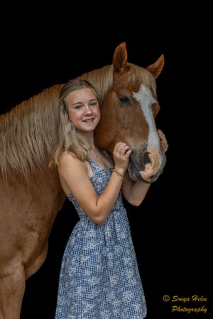 A person in a blue sundress smiles while posing with a chestnut horse against a dark backdrop.