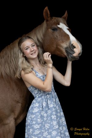 A person in a blue patterned dress poses playfully with a brown horse against a dark backdrop.