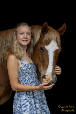 A person wearing a blue floral dress poses with a chestnut horse with distinctive white face markings.