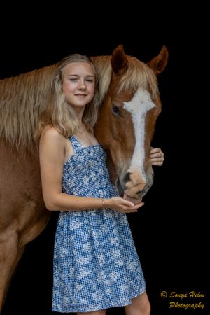 A person wearing a blue summer dress stands beside a chestnut horse with a white blaze.