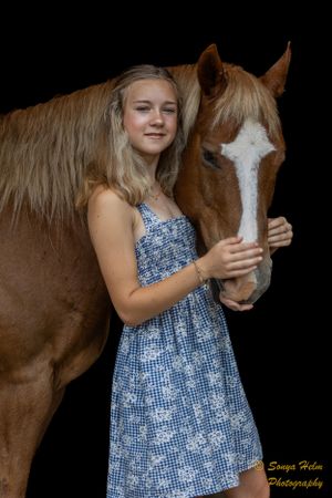 A person in a blue patterned dress stands with a chestnut horse against a dark background.