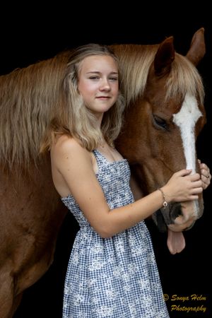 A person wearing a blue summer dress poses with a brown horse with a white blaze marking.