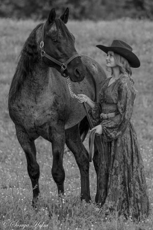 A black and white portrait of a horse and companion in vintage western attire against a rustic backdrop.