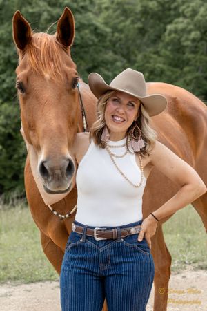 A chestnut horse and cowgirl in western attire pose together in a grassy field.