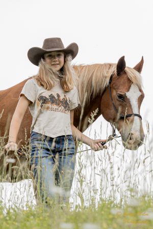 Person in graphic tee and cowboy hat stands with chestnut horse in wildflower field.