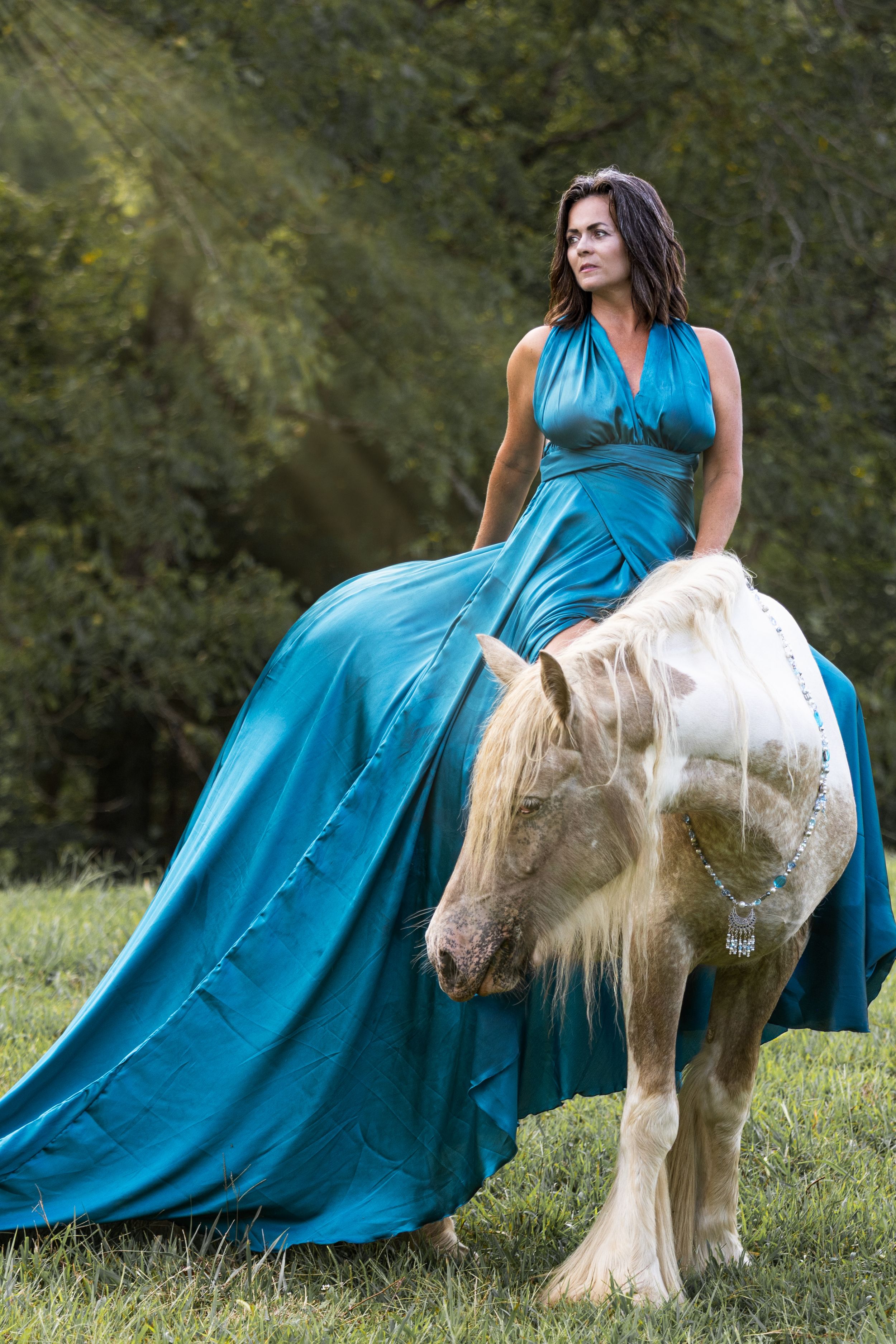 A white miniature horse stands beside someone in a turquoise gown in a sunlit meadow setting.