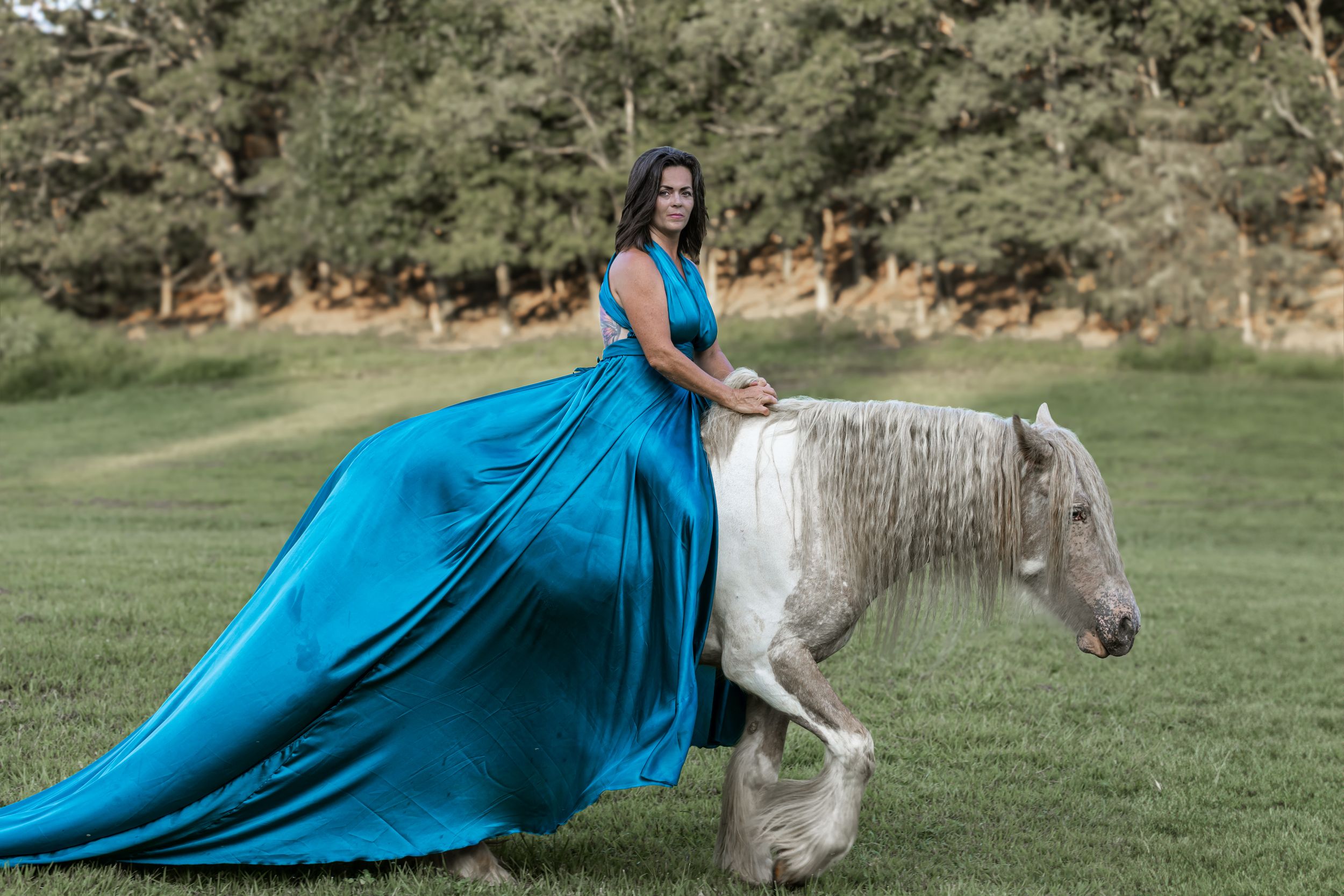 A person in a flowing turquoise gown poses elegantly with a white horse in a scenic grassy field.