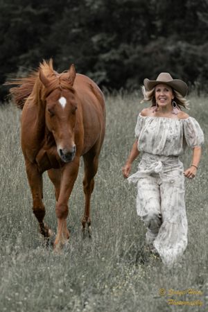 A chestnut horse and person in white dress run together through a grassy field.