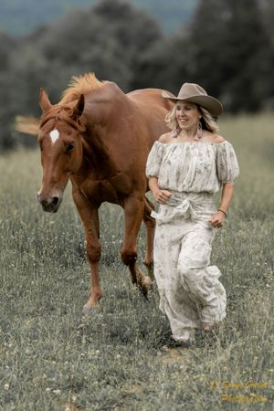 Person in white dress and cowboy hat walks alongside chestnut horse in meadow.