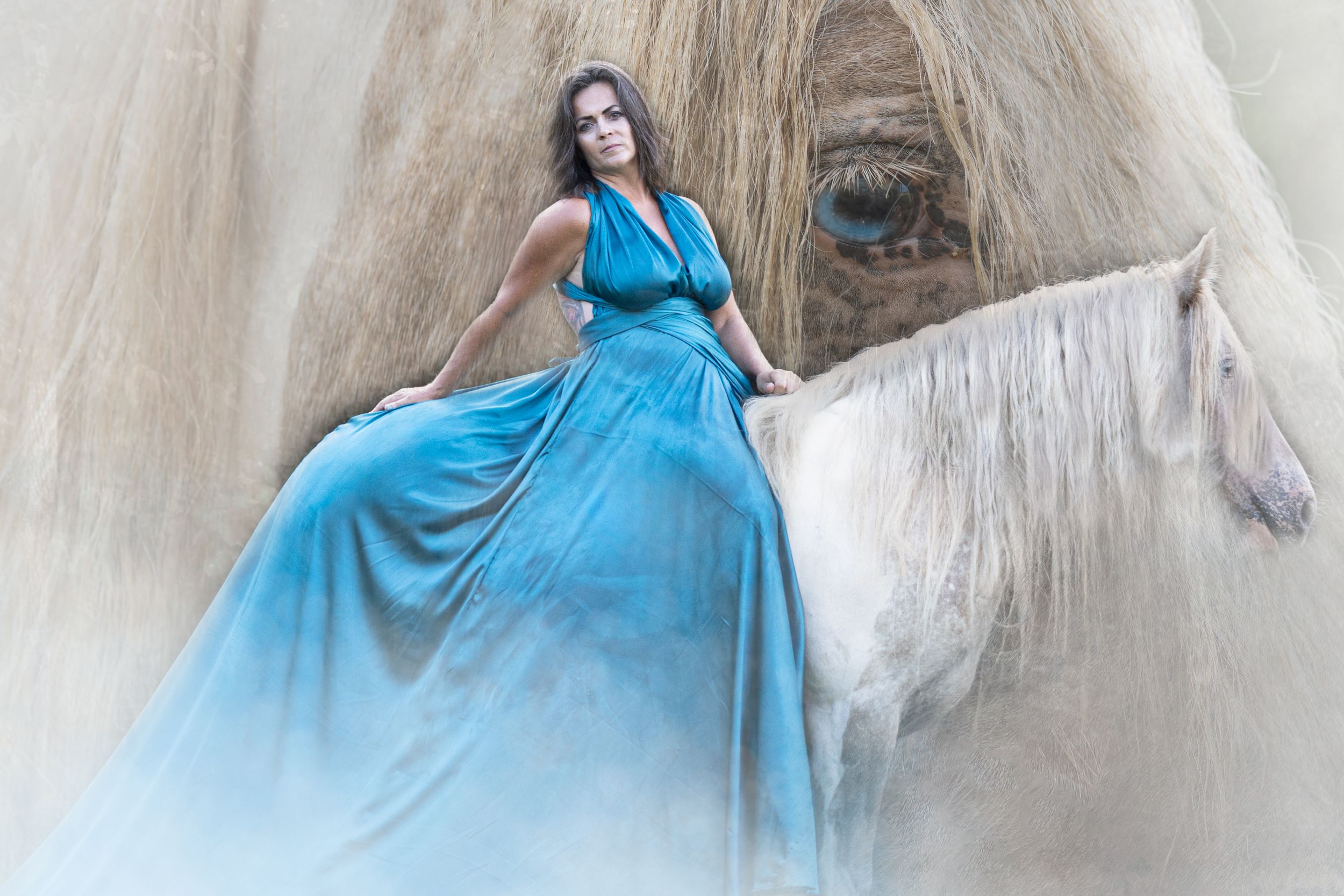 A woman in a flowing turquoise dress poses elegantly atop a majestic white horse in a dreamy setting.