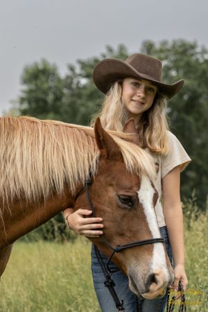 A brown horse with a white blaze nuzzles a cowgirl wearing a brown hat and white shirt in a sunny field.