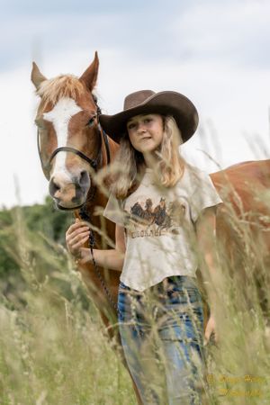 Person in western attire and cowboy hat poses with chestnut horse in tall grass.