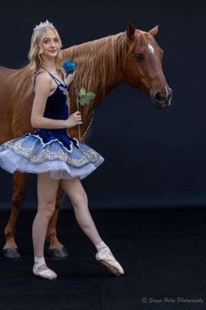 Ballerina in blue costume poses with flower beside chestnut horse against dark background.