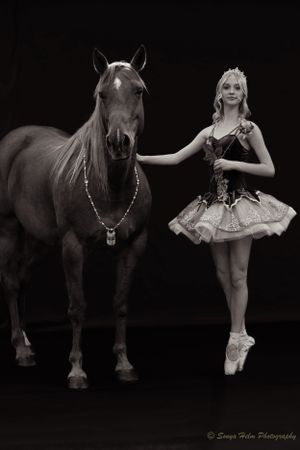 Monochrome studio portrait of classical ballerina posing beside adorned horse.