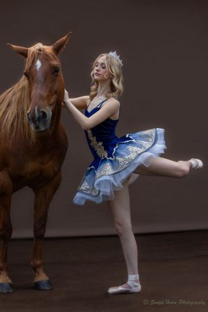 A ballet dancer in a blue tutu dress poses gracefully next to a chestnut horse against a dark backdrop.