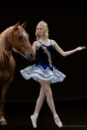 A dancer in a blue and white tutu performs a ballet pose beside a brown horse in a dark studio.