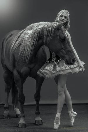 A black and white portrait of a ballet dancer posing with a horse in a studio setting.