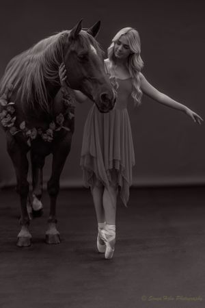 A dreamy black and white image of a dancer en pointe beside a horse wearing flowers.