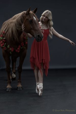 A ballerina in a red dress performs en pointe beside a horse wearing a floral garland.