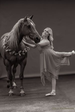 Ballet dancer in white dress poses gracefully beside a horse wearing a floral garland in black and white portrait.