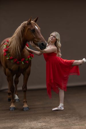 A ballerina in red performs an arabesque while touching a horse adorned with red flowers.