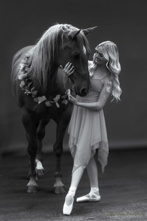 Elegant black and white portrait of dancer interacting with adorned horse in studio setting.