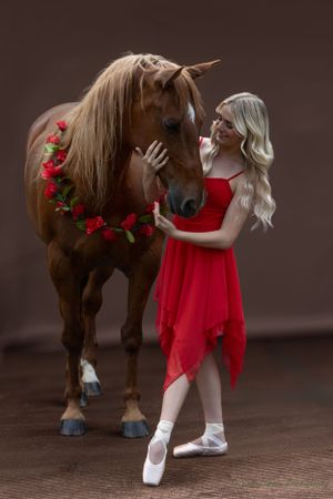 Ballerina in red dress poses with brown horse wearing rose garland against dark background.