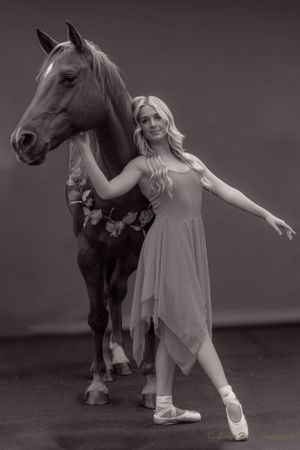 Black and white photo of dancer in flowing dress striking graceful pose beside decorated horse.