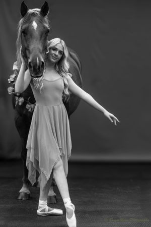 Monochrome image of ballerina in flowing dress posing elegantly with horse in studio setting.