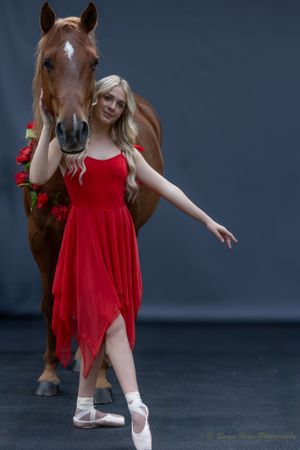 Dancer in red asymmetrical dress strikes ballet pose beside brown horse with rose garland.