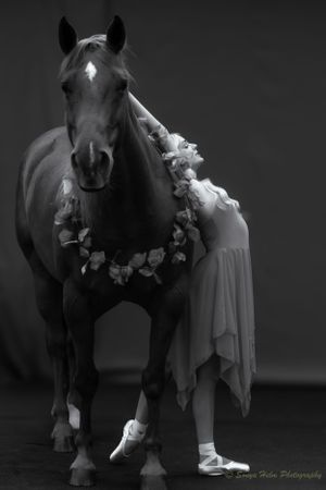 Artistic black and white photo of dancer in flowing dress beside horse wearing floral garland.