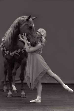 A graceful ballet dancer in a flowing dress poses with a majestic horse adorned with flowers in a black and white portrait.