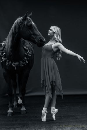 A ballet dancer en pointe strikes an elegant pose beside a dark horse wearing a floral garland.