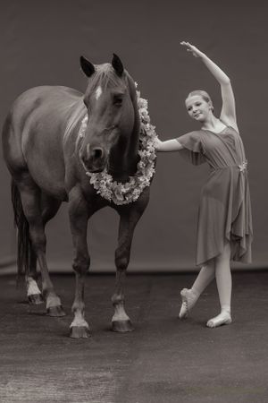 An artistic black and white portrait of a ballet dancer performing beside a flower-adorned horse.