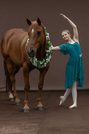 A ballet dancer in teal performs an elegant pose next to a horse wearing a floral wreath.