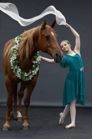 A ballerina in teal manipulates flowing white fabric while posing with a horse wearing flowers.