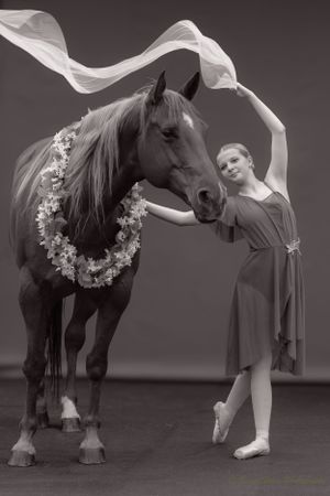 A monochrome portrait featuring a dancer with flowing fabric beside a horse wearing a floral wreath.