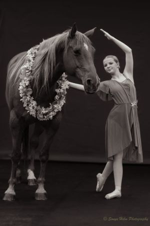 A dramatic black and white photo of a dancer posing with a horse wearing a floral garland.