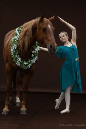 A ballet dancer in a teal dress performs gracefully next to a chestnut horse adorned with white flowers.