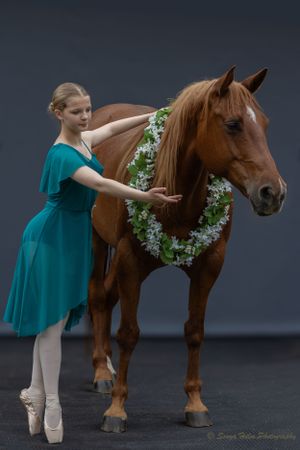 Ballerina in teal dress poses with horse wearing floral wreath.